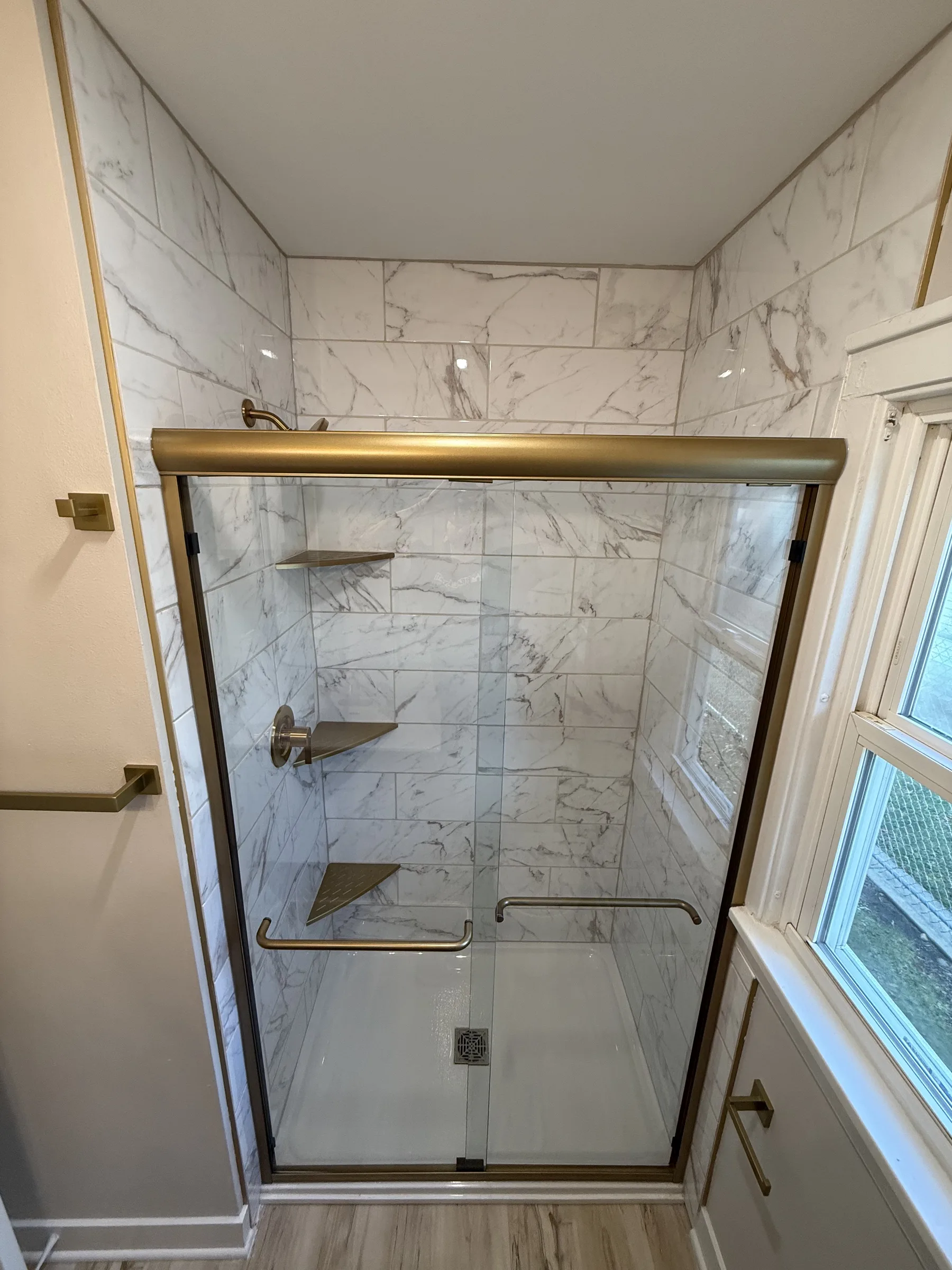 Marble-look tile walk-in shower with brass-framed sliding glass doors and tiled corner shelves, completed by NextGen Bathroom Specialists