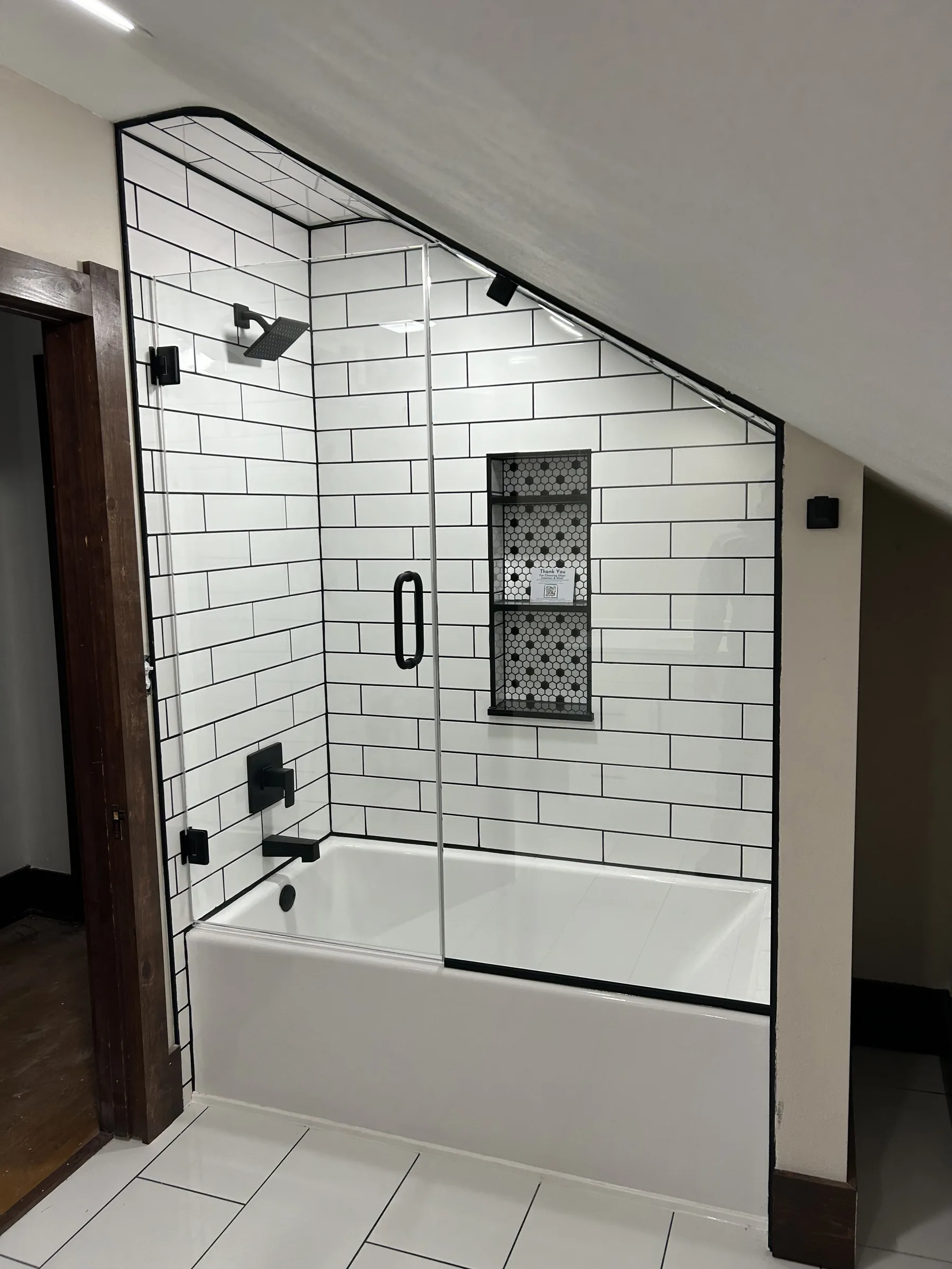 White subway tile tub and shower with black grout, matte black fixtures and a black-framed glass panel under a sloped ceiling, completed by NextGen Bathroom Specialists
