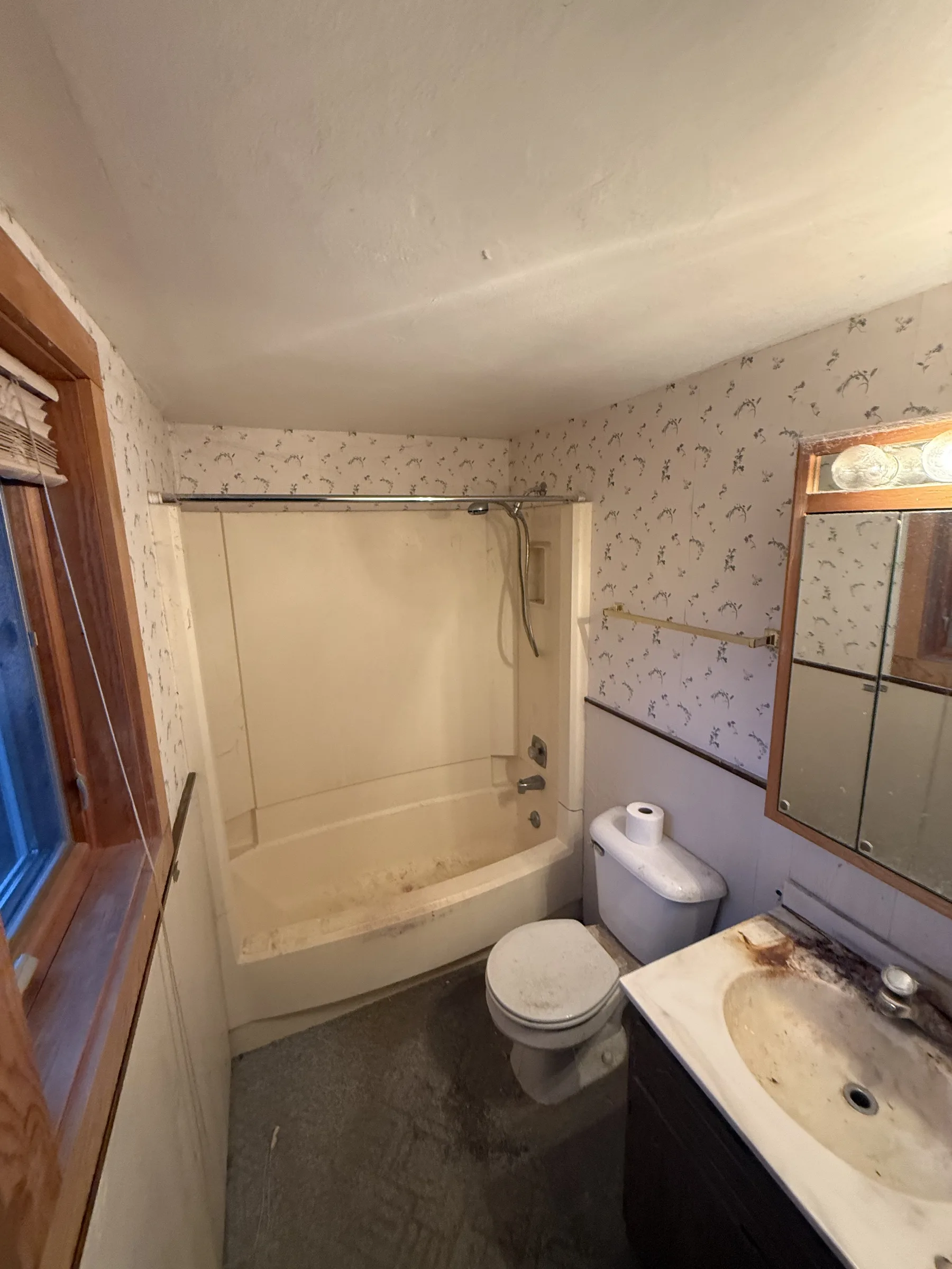Dated bathroom before renovation with floral wallpaper, a worn one-piece tub surround and a stained vanity sink